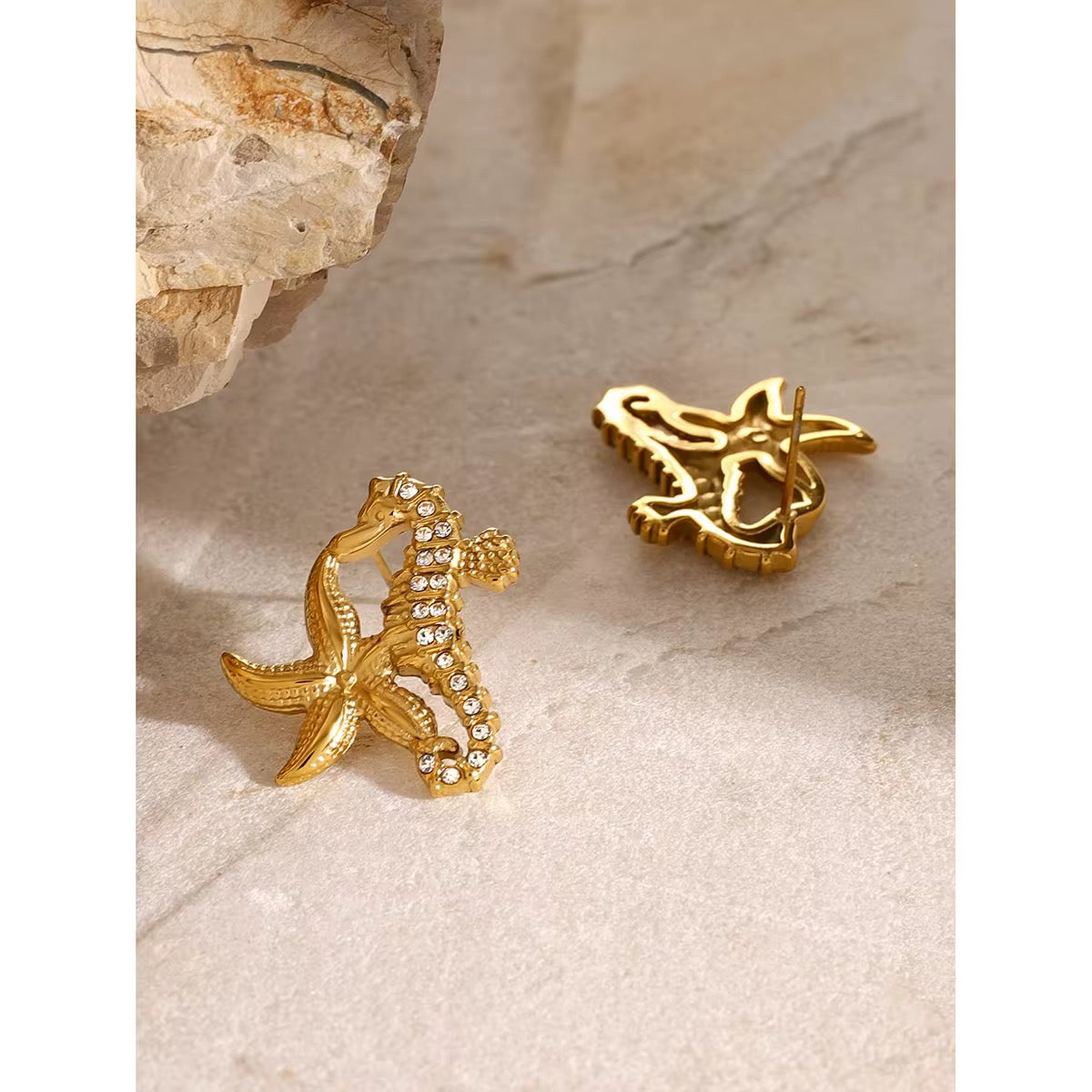 Ein Paar vergoldete Ohrringe aus Edelstahl in Form von Seepferdchen, präsentiert vor einem Stoffhintergrund.